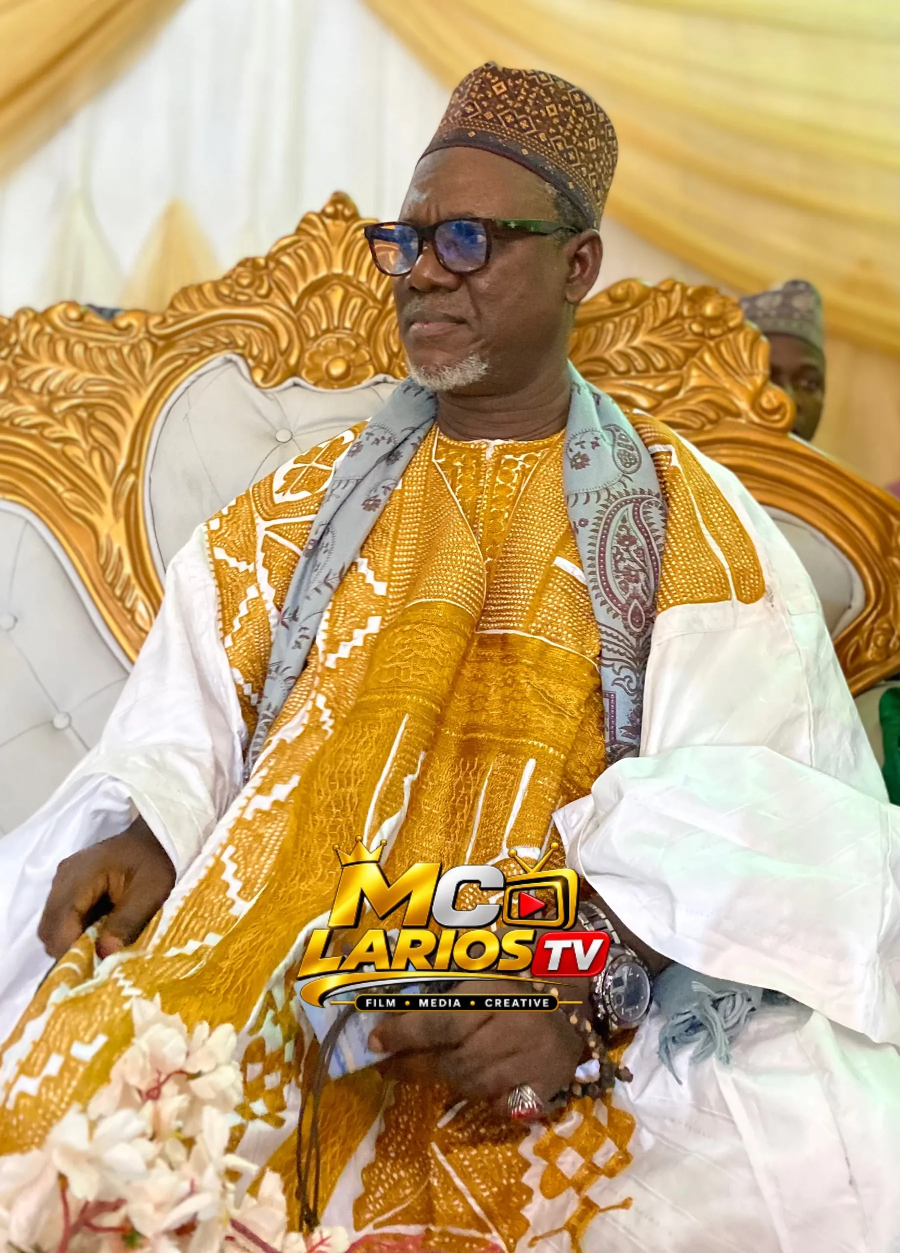 A dignitary in a white robe with gold embroidery and a patterned cap, seated on an ornate gold chair