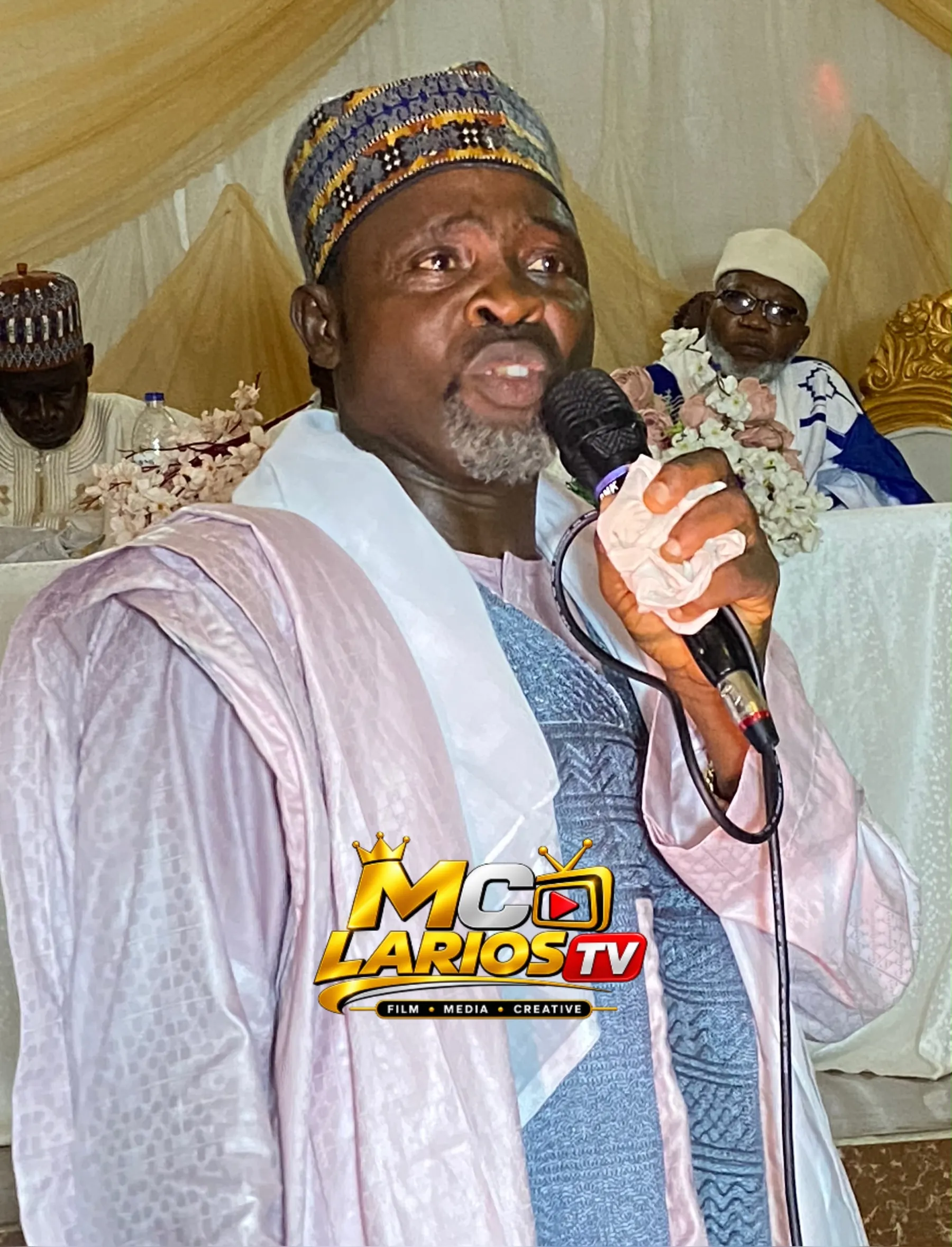 A man in a lilac robe and patterned cap speaking into a microphone