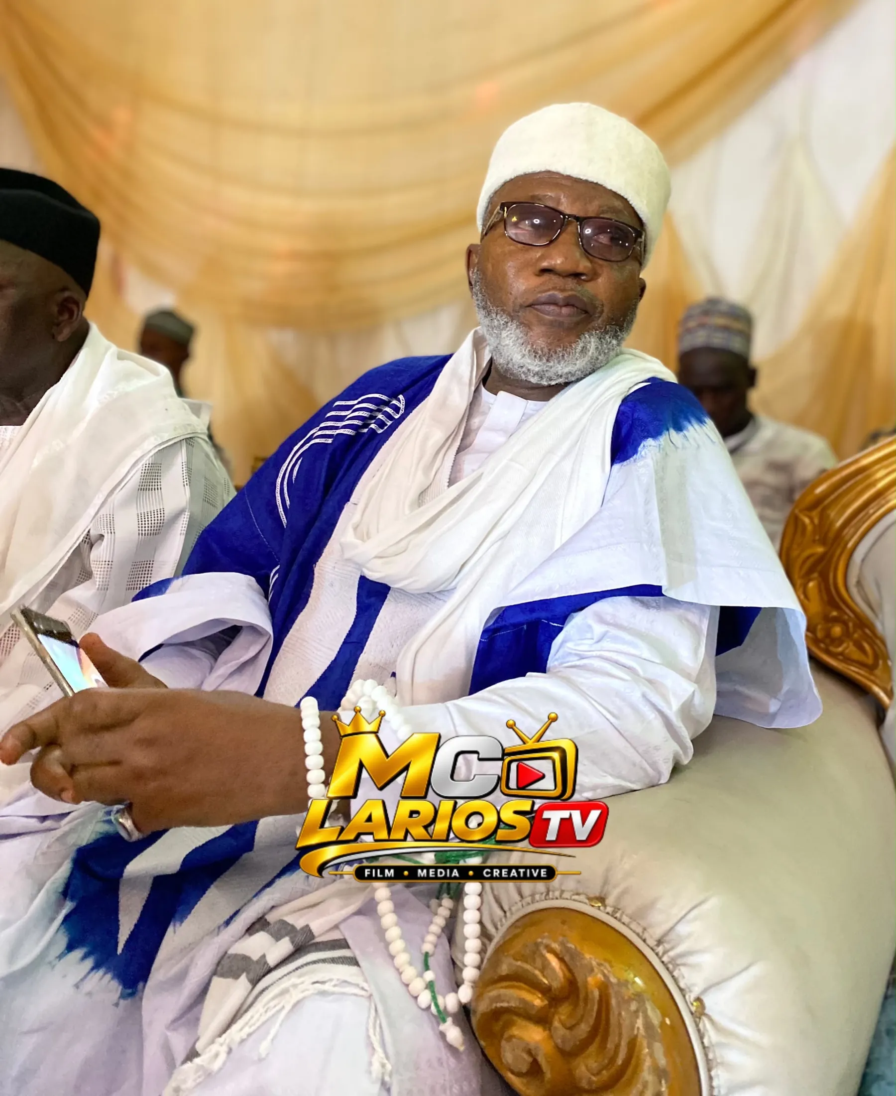 An elder in a white cap and blue and white robe, seated with prayer beads