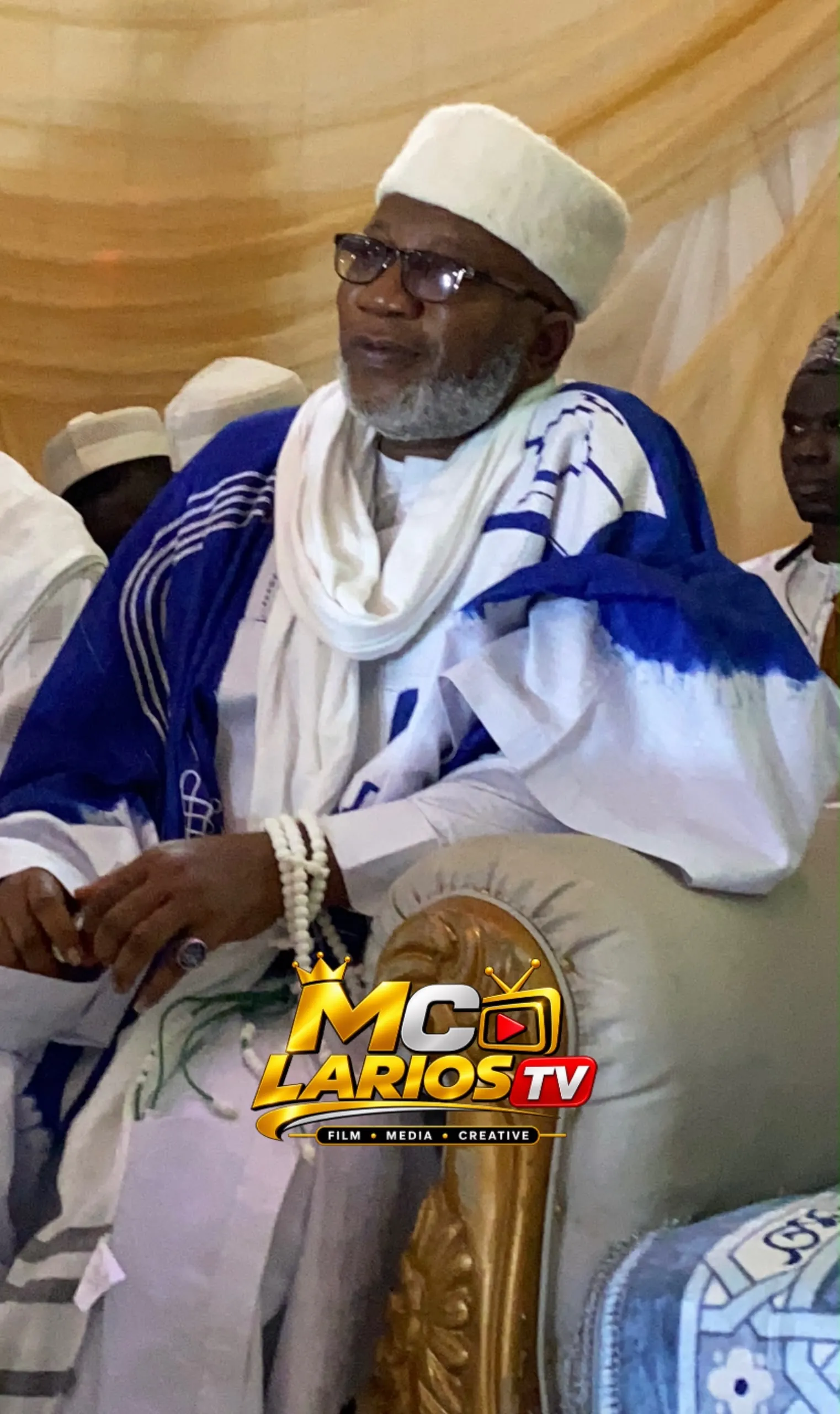 An elder in a white cap and blue and white robe seated on the high table