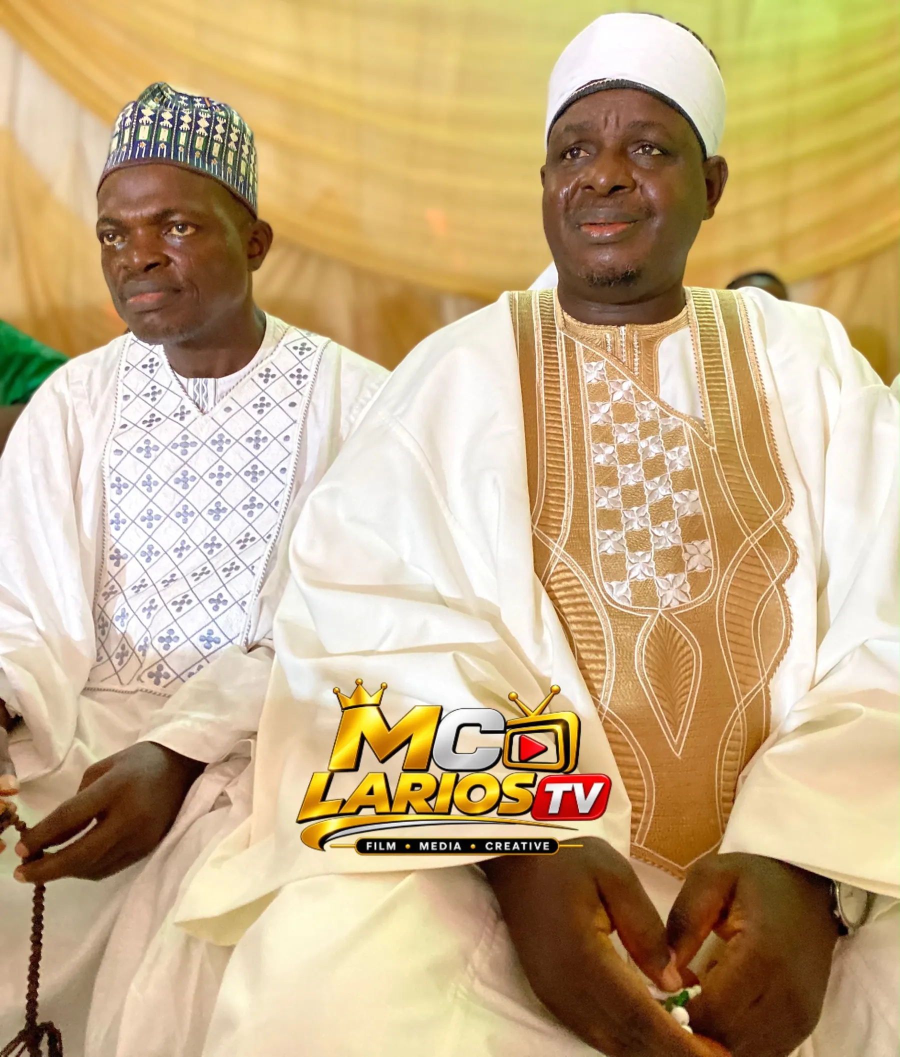 Two men in white robes seated side by side, one holding prayer beads