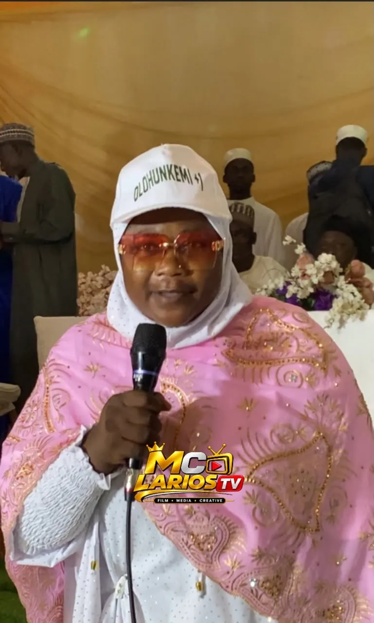 A woman in a white cap and pink shawl speaking into a microphone